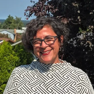 Amara, una mujer de mediana edad, de tez morena clara y cabello castaño corto y rizado, sonríe afuera. Viste una camisa blanca con diseños geométricos y gafas con montura negra. 