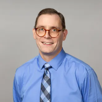 Un retrato de Robert Stoll, sonriendo y con gafas redondas, camisa azul y corbata a cuadros.