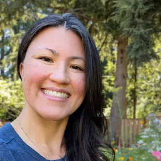 Ellen smiles at the camera, she has long dark hair and is wearing a blue shirt in front of a forested background. 