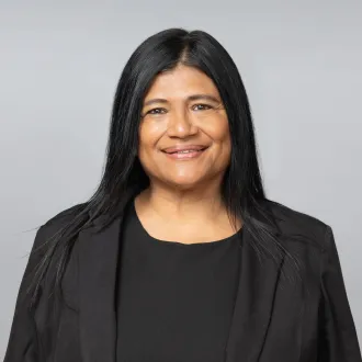 Retrato de Irma Jiménez. Aparece sonriendo, con cabello largo, liso y negro. Viste una blusa y un blazer negros sobre un fondo gris claro.