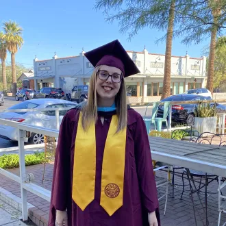 Alyssa, a light skinned woman with long blond hair and glasses, smiles outdoors wearing a maroon graduation gown