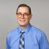 A headshot of Robert Stoll, smiling and wearing round glasses, a blue shirt, and a checkered tie.