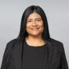 A headshot of Irma Jimenez. She is smiling and has long, straight black hair with black top and blazer against a solid light gray background.