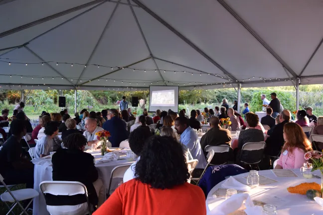 Over 150 people seated at tables under a large white tent watching a video.