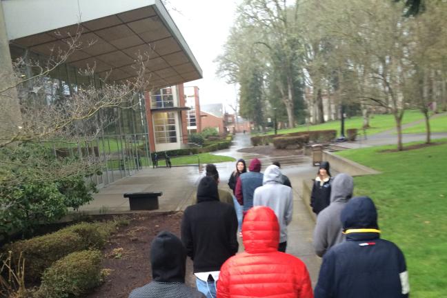 A group of young adults walk away from the camera on a sidewalk in the rain. 