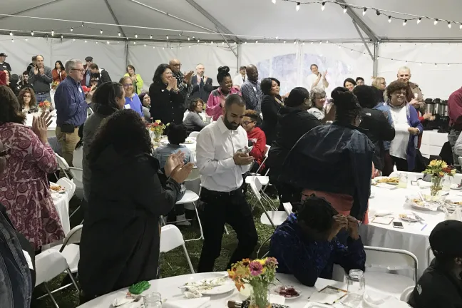A large group of people stand around tables under a tent applauding and smiling.