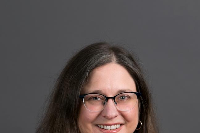 Formal portrait of Auditor McGuirk shown with long brown hair, glasses and wide smile.