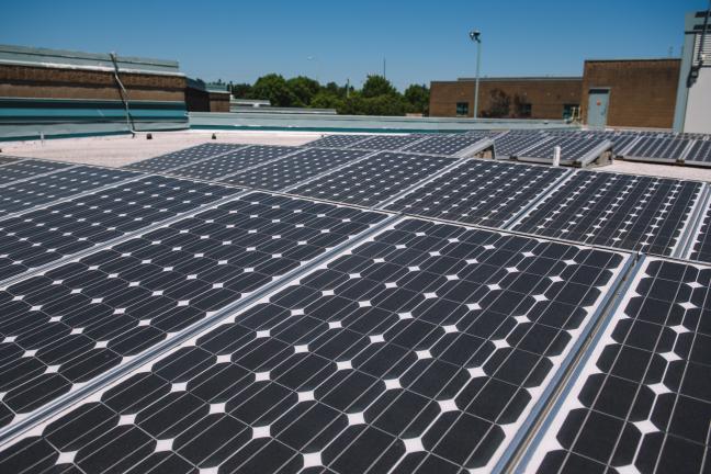 Picture of solar panels on a roof, blue sky overhead, trees in the distance.