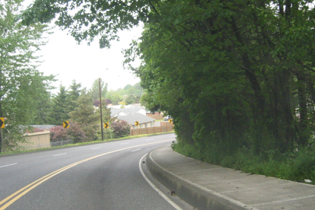 A driver's-eye view of a curve in the road, with green trees on one side and houses at the bottom of a hill on the other.