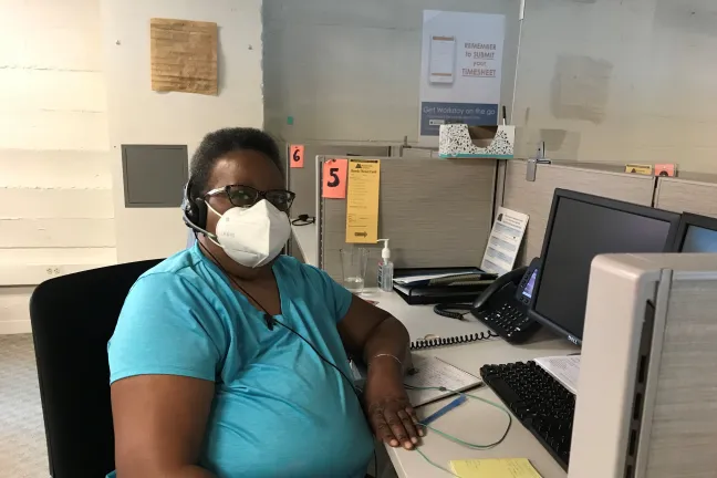 Delores Terry, election worker sits at her station in the elections call center. She wears a white face covering and blue shirt. A copy of the constitution hangs on the wall behind her.