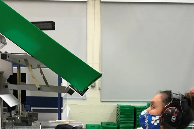 Kristen Mun, elections worker stands at the sorter machine. She takes ballots from a tray at shoulder level and puts them into a green tray just above her head. She wears black headphones, a blue mask with white flowers on it, and a cream colored shirt.