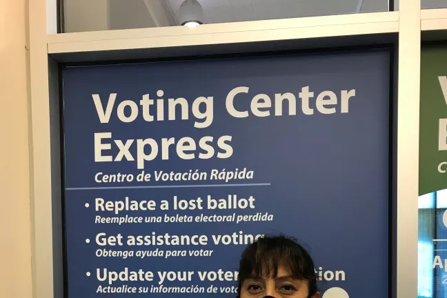Susy Kristin, elections worker poses in front of the Gresham Voting Center Express bilingual sign in Gresham. She wears a black face covering, a pink shirt, and white sweater. She has a badge pinned to her shirt that says Habla Español.