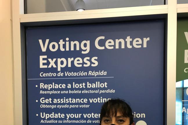 Susy Kristin, elections worker poses in front of the Gresham Voting Center Express bilingual sign in Gresham. She wears a black face covering, a pink shirt, and white sweater. She has a badge pinned to her shirt that says Habla Español. 