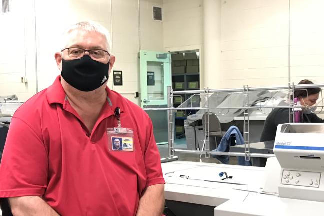 Tom Urbanowicz, election worker sits at a ballot extration table. He wears a black face covering and red colored shirt. There is a woman working at a machine behind him and an open green door in the background.