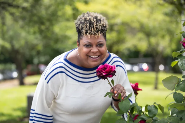 Cynthia Lambert in white and blue top, bending down to smell a rose
