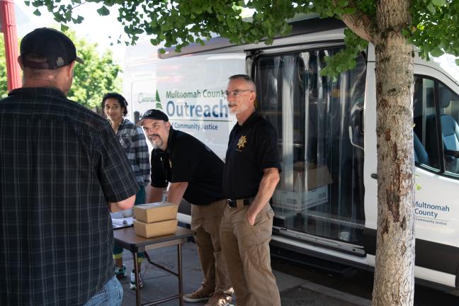 Mental Health Unit officers John McVay and Joe Wenhold, alongside College to County intern Zola Neal, set up a stand and lay out socks, bottles of water and box lunches