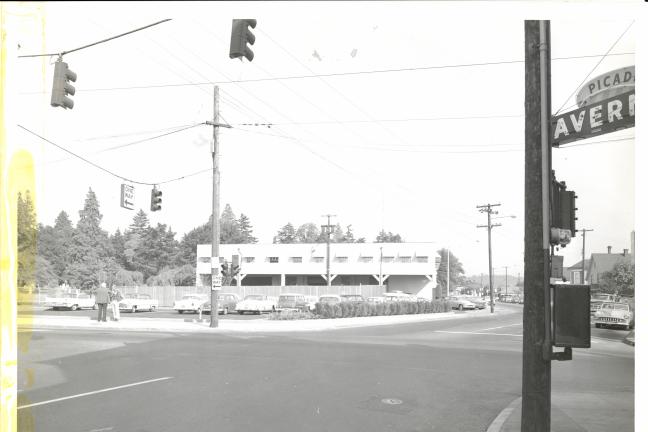 Photograph looking diagonally across a stoplight intersection at a nondescript two-story office building. Two men stand talking on the sidewalk next to a small parking lot with several vehicles.