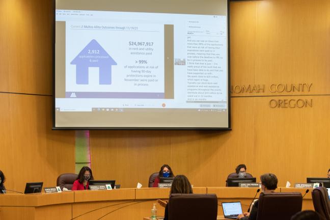 The five commissioners listen to two presenters during a briefing about rent assistance in the boardroom. The screen above the commissioners shows a slide from the presentation.