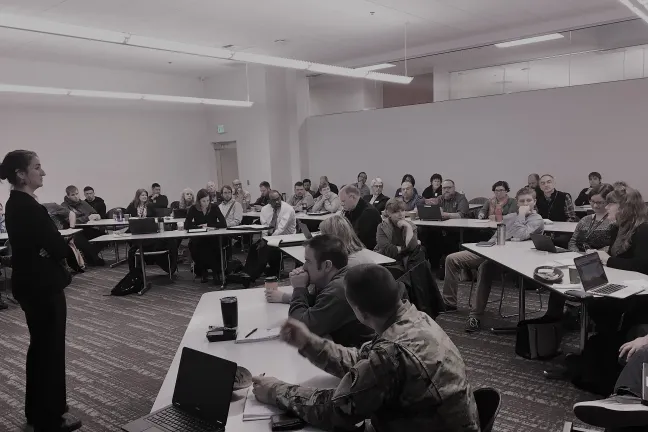 Group of people sitting at tables in a building listening to a woman presenter 