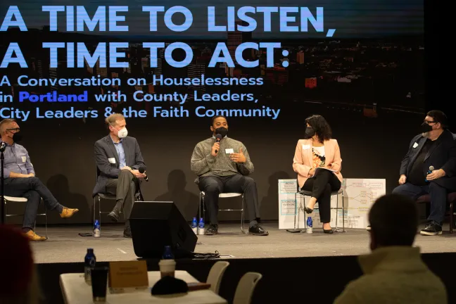 Six people sitting on stage. A projection behind them reads "A Time to Listen, A Time to Act"