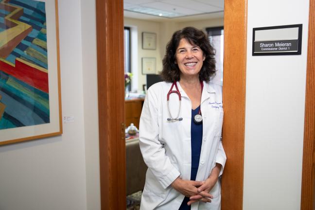 Commissioner Sharon Meieran standing in the oak doorway of her office wearing her white doctor's coat and stethoscope. 