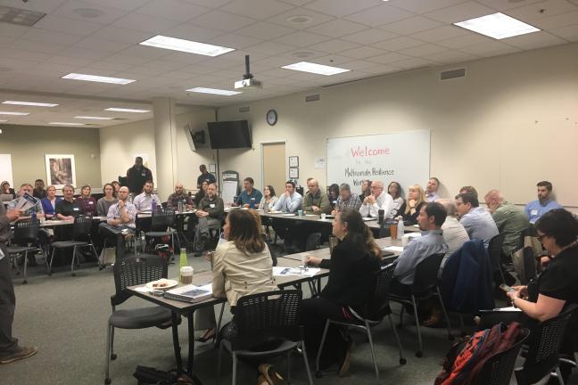 Image of many people sitting in a room in a horseshoe shape listening to a lecure