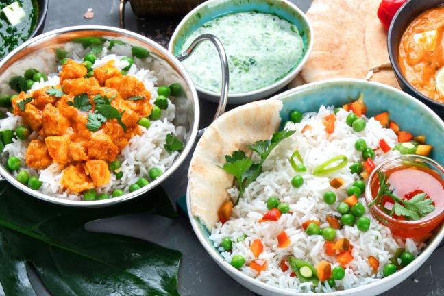 A variety of different foods on a table, including rice topped with vegetables, samosas, curries, and chicken drumsticks