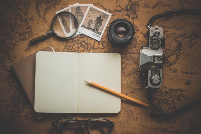 An SLR camera, magnifying glass, Polaroid photographs, a pair of glasses, and a journal open to a blank page with a pencil 