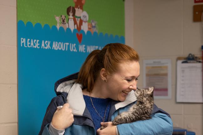 A woman in a blue jacket holds a gray and white kitten up to her face, gently touching its nose to hers.