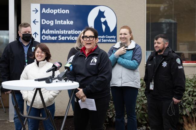 Six people lined up behind a stand with several microphones propped on it. MCAS Director Erin Grahek stands directly behind the microphones and speaks.