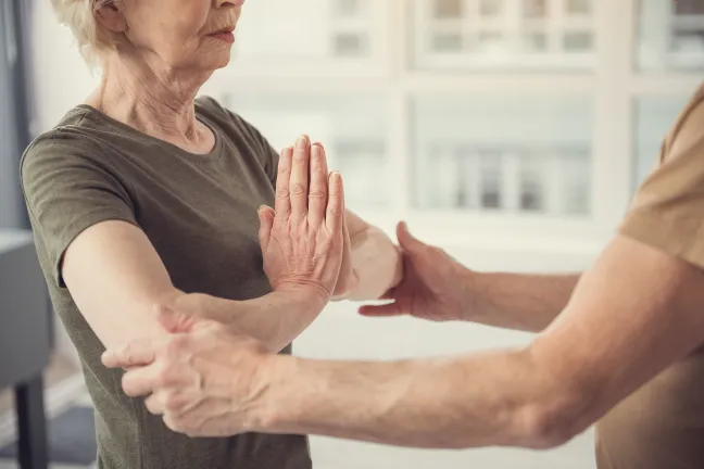An older adult woman with white hair and a green shirt standing with her palms pressed together. An instructor reaches out to adjust the position of her elbows.