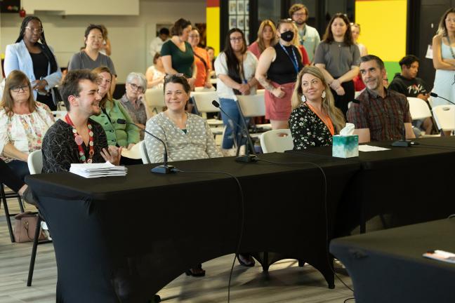 Four people sitting behind a table draped in a black tablecloth; the individual on the left, with a stack of paper in front of them, is smiling and speaking; the other three are looking at them; many people in the audience behind them are standing.