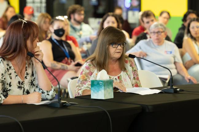 A woman wearing glasses sit at a table draped in black tablecloth, speaking into a microphone.