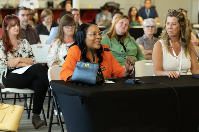 Former County Commissioner Loretta Smith sits behind a a table draped in a black tablecloth, smiling and holding the stem of a microphone.