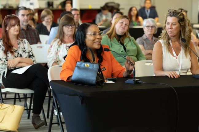 Former County Commissioner Loretta Smith sits behind a a table draped in a black tablecloth, smiling and holding the stem of a microphone.