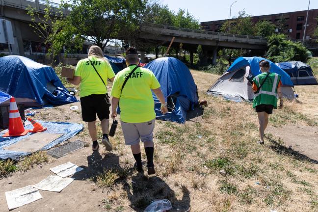 A group of outreach workers walk toward tents set up on grass under a bridge.