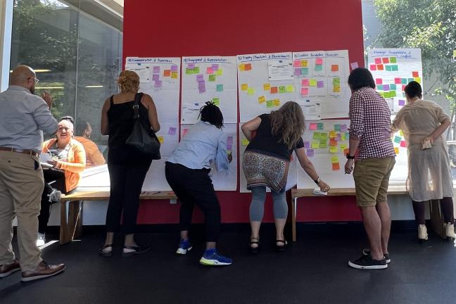 Five people facing a wall: several are looking at large sheets of paper on a red wall and a window with sticky notes on them; others are in adding sticky notes to the paper; to the left, a standing person talks to a seated person