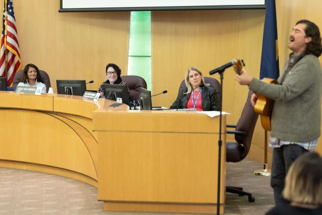 Photo of Board of Commissioners watching as local artist Joaquin Lopez performs with guitar