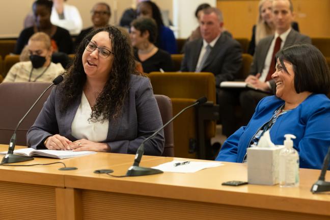 Rachael Banks sits at a table addressing an audience off camera. She sits behind a microphone on the table with a notebook placed in front of her. Chief Operating Officer Serena Cruz sits, smiling, to Rachael's left.