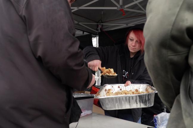 A woman wearing a sweatshirt serves food to people standing outside below a tent.