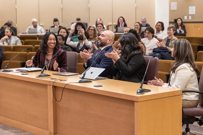 In the foreground, four people sit behind a large table celebrating with applause. In the background is a seated audience looking happy.