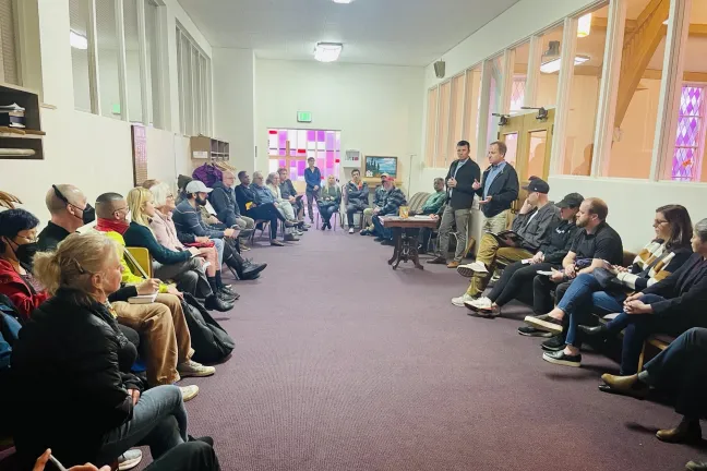 A large group of people sitting in a hallway listen to two speakers who are standing.