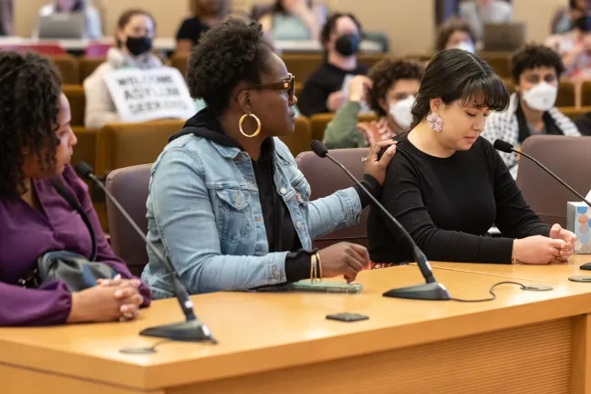 Three people sit at dais in Multnomah County Boardroom. One person places hand on the shoulder of the person sitting next to them.