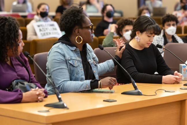 Three people sit at dais in Multnomah County Boardroom. One person places hand on the shoulder of the person sitting next to them.