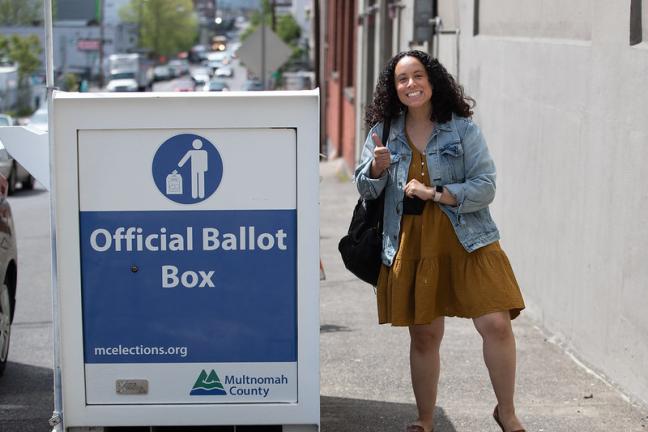 Person with the thumbs up hand right signal standing next to Multnomah County elections official ballot box with signage that reads official ballot box