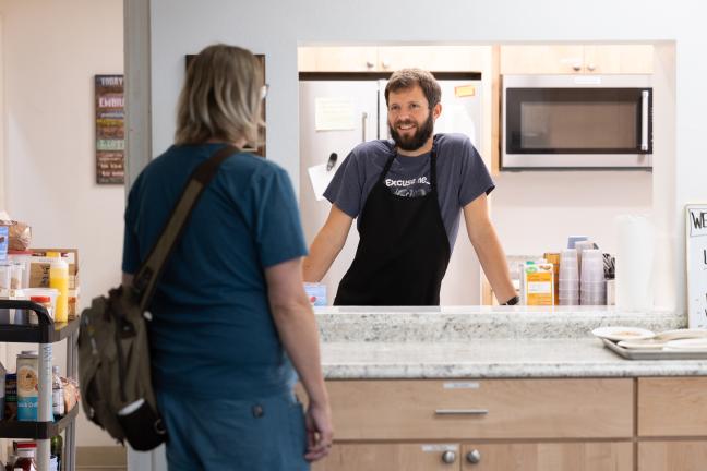 A person inside a kitchen wearing an apron talks through a window to another person.