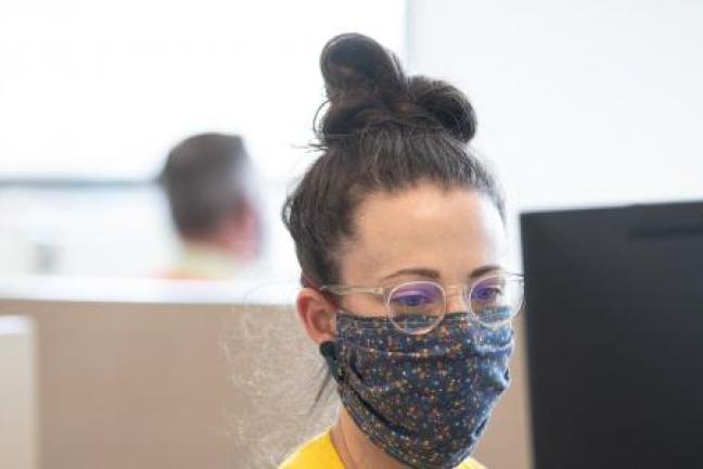 Multnomah County staff person with brown hair in a bun and glasses with mask on sits at a computer at the Emergency Operations Center