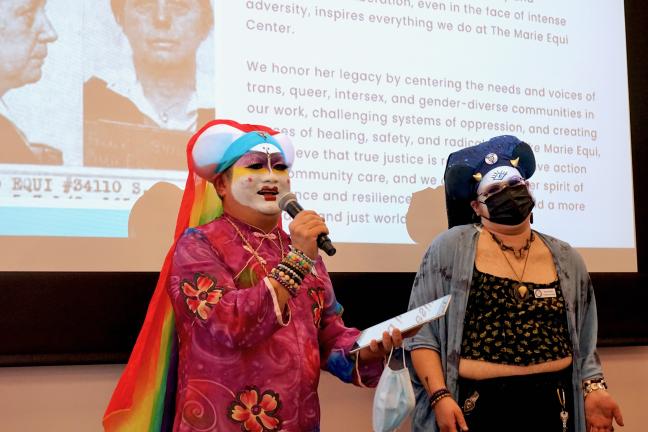 Two people wearing theatrical makeup and head coverings talk in front of a projector.