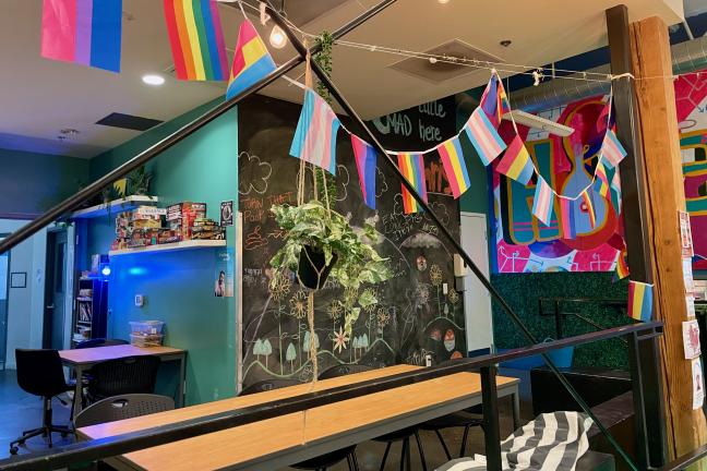 Pride flags hang on a garland over a room with tables and a chalkboard wall.