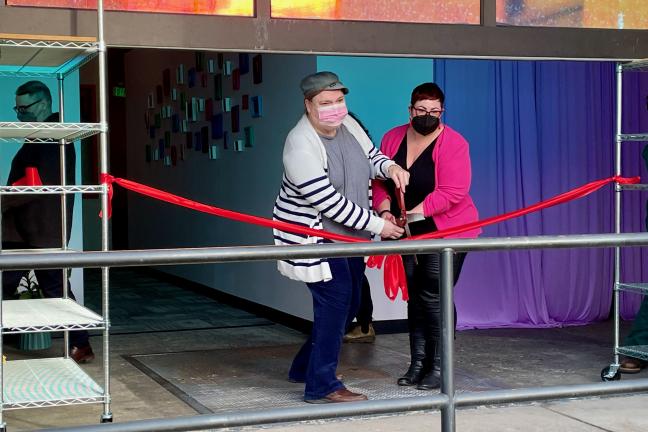 Two people hold up big scissors, cutting ribbon in front of a building.